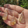 Hand holding three gold kuan yin rutilated quartz carved necklaces against a green leafy background