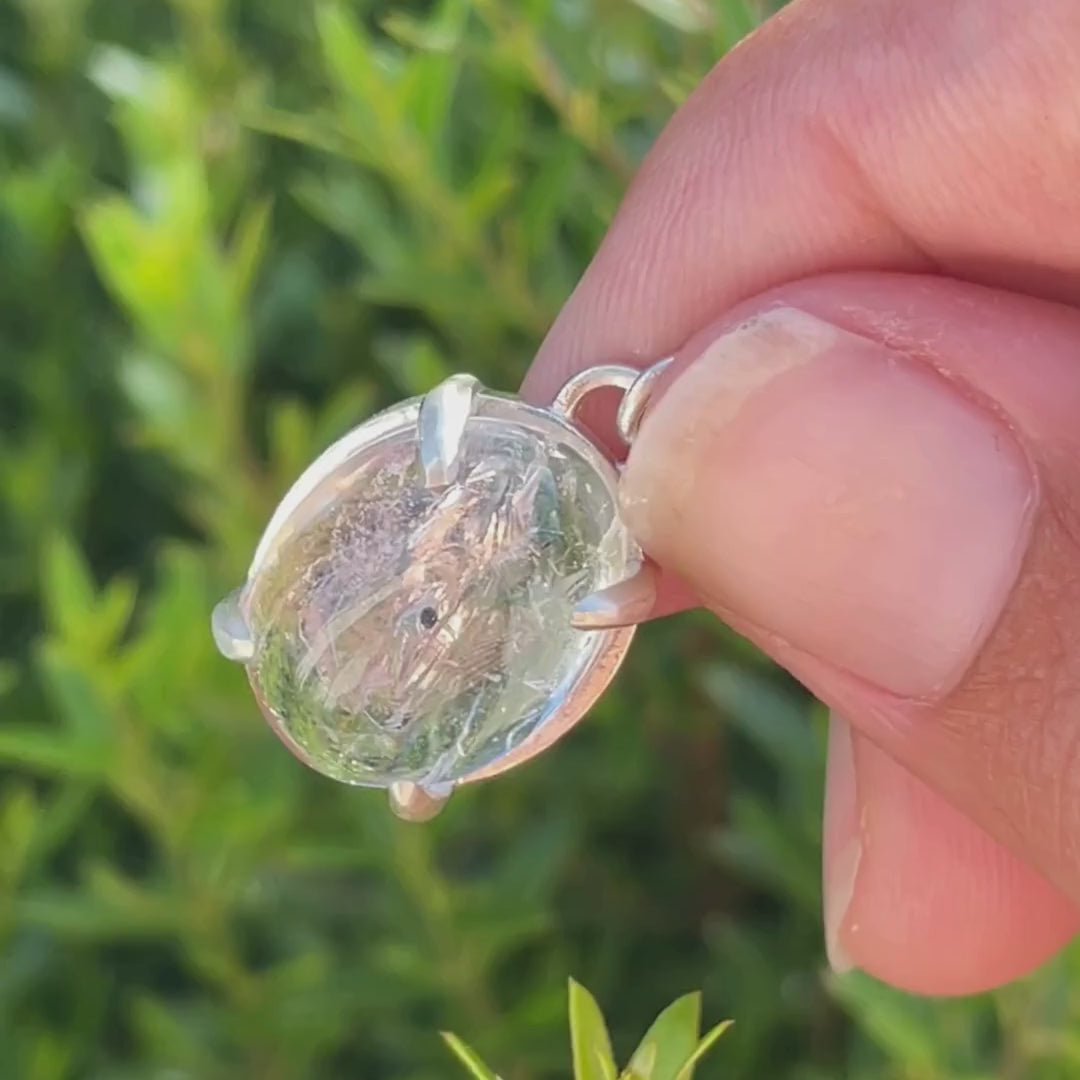 Silver necklace with a enhydro quartz pendant held between fingers against a green leafy background moving to show bubble.